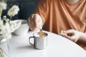 Cropped view of unrecognizable woman in a tangerine top lighting a candle inside a metal mug,