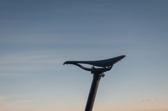 A minimalist silhouette of a bicycle saddle seat set against a soft sunset sky. The image evokes a