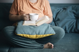 Cropped unrecognizable woman sits comfortably on a couch with a book and a mug of tea, embracing a
