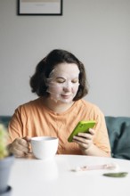 Woman with a facial mask resting at home while holding a cup of coffee and browsing on her