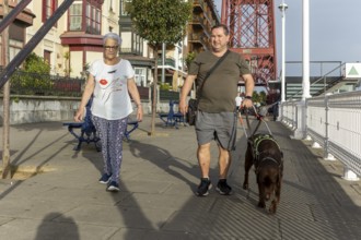 A blind man and senior woman walks their guide dog along a sunny urban street with a prominent