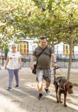 A blind man with a guide dog strolls through a sunlit park, accompanied by a woman. The scene