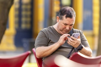 A blind man sitting on a red chair outdoors, using his smartphone. The background features muted