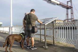 A blind man with a guide dog read an information board on a bridge overlooking a scenic river and