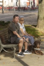 A blind man and an elderly woman sit on a park bench, enjoying the outdoors with a guide dog by