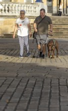 A blind man and senior woman walks together on a sunny day, accompanied by a guide dog. They stroll