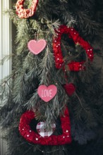 Valentine's Day ornaments in heart shapes hang from evergreen branches, featuring red and pink hues
