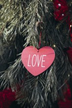 A red heart-shaped ornament with the word LOVE hanging among lush green pine tree branches,