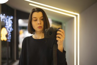 A woman capturing a self-portrait using a DSLR camera in a well-lit interior with neon accents and