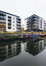 A modern London cityscape features sleek apartment buildings and colorful canal boats reflected in