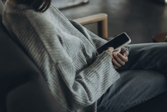 A person wearing a comfy sweater and jeans relaxes on a sofa, holding a smartphone. The setting