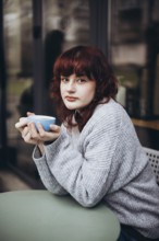 A young woman with curly red hair relaxes at a cafe, dressed in a cozy sweater. She holds a