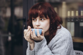 A young woman with red hair sips from a blue cup of coffee, wrapped in a cozy sweater. She gazes