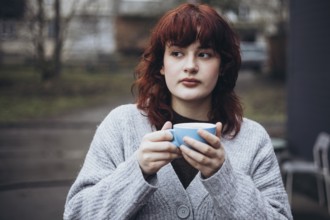 A young woman with red hair, wearing a grey sweater, holds a blue mug of coffee in an outdoor urban