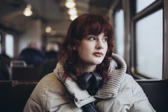 A young woman with headphones, wearing a warm jacket, gazes thoughtfully out the window of a train,