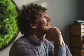 A curly-haired young man smiles contemplatively, sitting against a vibrant green wall. The imagery