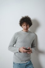 A curly-haired young man leans casually against a plain wall, focused on his smartphone. He wears a