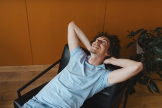 A curly-haired young man in a light blue shirt, lounging back in a modern chair with an orange