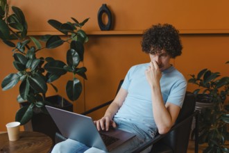A curly-haired young man in a light blue shirt works on his laptop in a stylish room with plants