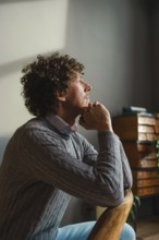 A contemplative man with curly hair sits by a window, resting his chin on his hands. Warm natural