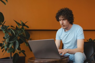 A curly-haired young man focused on his laptop, sitting in a modern space with orange walls and