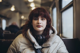 A young woman with headphones rests on a train seat wearing a warm jacket. The blurred background