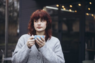 A young woman with red hair, wearing a gray cardigan, holding a coffee cup and gazing thoughtfully