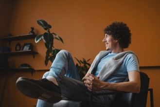 A curly-haired young man sits comfortably in a modern living room with a warm orange background,