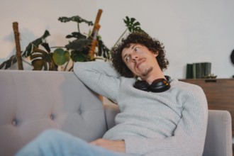 A curly-haired young man with casual attire, relaxing on a sofa with headphones around his neck.