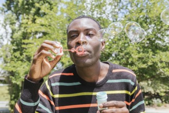 A man in a striped sweater joyfully blows bubbles outside on a bright sunny day, surrounded by lush