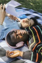 Friends relax outdoors on a sunny day, lying on a blanket. One listens to music with headphones,