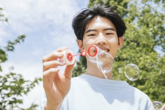 A smiling man enjoys blowing soap bubbles in a sunlit park. The clear sky and vibrant greenery