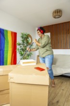 A trans woman smiling while organizing her new home. Surrounded by moving boxes and a pride flag,