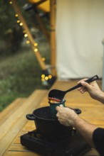 Cropped unrecognizable person pours spicy mulled wine into a mug, outdoors near twinkling lights