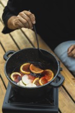 Cropped unrecognizable person stirs a pot of mulled wine on an outdoor burner, featuring sliced