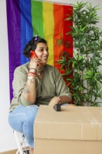 Trans woman sitting by moving boxes, talking on phone, smiling. Vibrant rainbow pride flag in