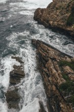 A dramatic aerial view of a rocky coastline where powerful ocean waves crash against rugged cliffs.