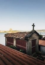 A traditional stone horreo granary with a cross atop its red tile roof overlooks a tranquil coastal