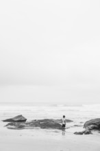 A lone individual stands on a beach surrounded by rocks, gazing at the ocean. The black and white