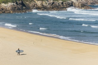 A surfer carrying a surfboard walks alone on a sandy beach toward the ocean, with waves crashing