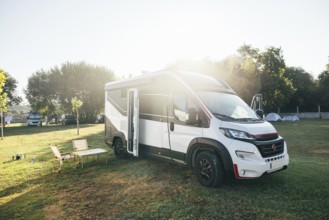 A modern camper van is parked in a sunny campground, complete with outdoor chairs and a table. The