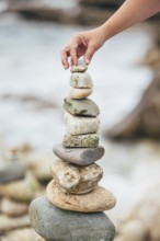 A hand carefully places a small stone atop a balanced stack of larger rocks by the seaside. The
