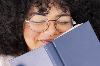 Close-up of a young latin woman with curly hair and glasses, smiling while reading a blue book. She