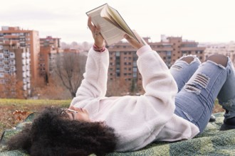 A woman lies on a blanket in a park, reading a book. They wear casual clothes, including jeans and