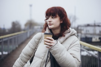 A young woman stands on a pedestrian bridge, wearing a warm jacket and holding a cup of coffee in a