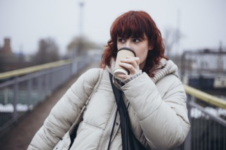 A young woman in a warm winter coat drinks coffee from a disposable cup in a day of travel by
