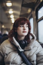 A young woman in a cozy winter coat sits on a train, gazing thoughtfully out the window. Headphones