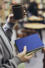 A woman in casual attire holds a coffee mug and books, enjoying a quiet moment in a cozy bookstore