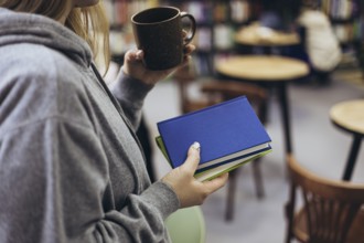 A woman in a gray hoodie holds books and a cup of coffee, enjoying a relaxed moment in a cozy