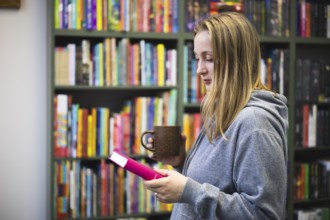 A woman in a gray hoodie enjoys a quiet moment in a bookstore. They hold a pink book and a brown